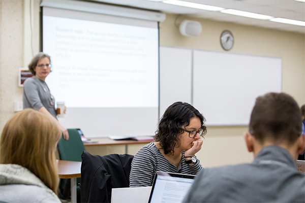 small lecture room with students in the foreground and professor giving powerpoint presentation